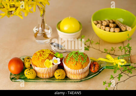 Muffins decorated with sprinkles, flowers of mastic, colored easter quail eggs on a kitchen table. Happy Easter holiday and greeting card concept. Stock Photo