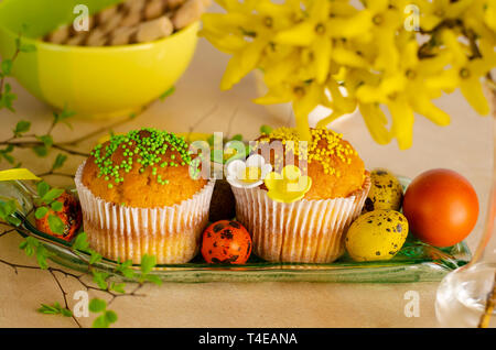 Muffins decorated with sprinkles, flowers of mastic, colored easter quail eggs on a kitchen table. Happy Easter holiday and greeting card concept. Stock Photo