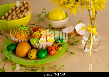 Muffins decorated with sprinkles, flowers of mastic, colored easter quail eggs on a green plateon the kitchen table. Happy Easter holiday and greeting Stock Photo