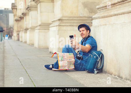 Smiling mature man using mobile phone while sitting against building column – happy tourist taking selfie with smartphone – traveler with backpack Stock Photo