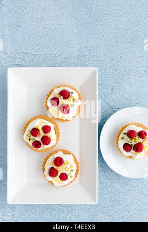 Homemade raspberry tartlets with whipped cream, chopped pistachio nuts and lemon zest on white porcelain serving plate. Top view. Stock Photo