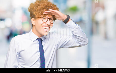 Young handsome business man with afro wearing glasses very happy and smiling looking far away with hand over head. Searching concept. Stock Photo
