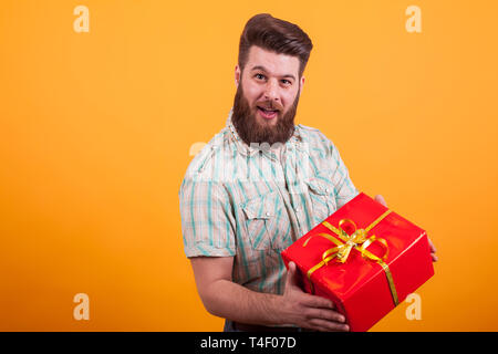 Bearded young man holding a red gift box and looking at the camera over yellow background. Cool dude.Attractive man. Stock Photo