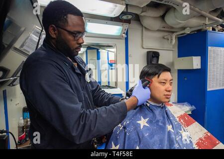 ATLANTIC OCEAN (April 6, 2019) Hospital Corpsman 3rd Class Nathaniel Li, right, from Gaithersburg, Md., receives a haircut from Ship’s Servicemen 3rd Class Malik Ball, from New Orleans, La., aboard the Ticonderoga-class guided-missile cruiser USS Leyte Gulf (CG 55). Leyte Gulf is underway as part of the Abraham Lincoln Carrier Strike Group (ABECSG) deployment in support of maritime security cooperation efforts in the U.S. 5th, 6th and 7th Fleet areas of responsibility. With Abraham Lincoln as the flagship, deployed strike group assets include staffs, ships and aircraft of Carrier Strike Group  Stock Photo