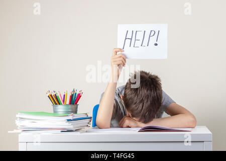 Sad tired frustrated boy sitting at the table with many books and holding paper with word Help. Learning difficulties, education concept Stock Photo