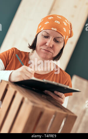 Female carpenter writing DIY project notes after finishing wooden crate in her small business woodwork workshop Stock Photo