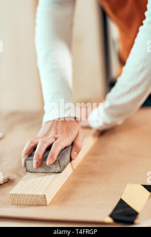 Female carpenter manually sanding wooden plank woth sponge sander in small business woodwork workshop, selective focus Stock Photo