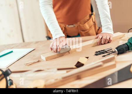 Female carpenter manually sanding wooden plank woth sponge sander in small business woodwork workshop, selective focus Stock Photo