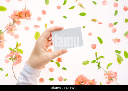 Woman holding blank white business card mock up in hand over white background with blooming springtime flower petals and leaves Stock Photo