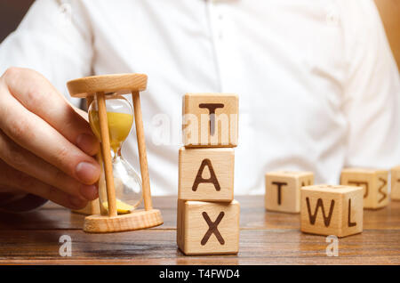 Businessman's hand holding a hourglass near the wooden blocks with the word Tax. Time to pay taxes. The concept of annual taxation. Taxes on vehicles, Stock Photo