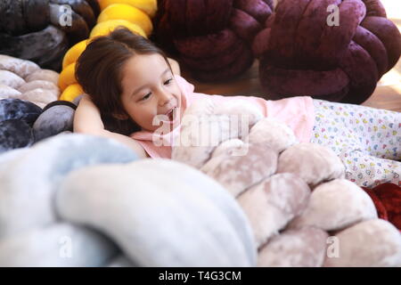 Child girl plays on the wooden floor among unusual multicolor pillows in pajamas. Closeup. Stock Photo