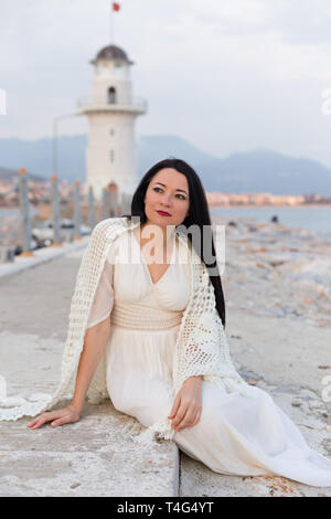 Brunette female dressed in boho style clothes sitting on the rocks with lighthouse and mountains blurred as background Stock Photo