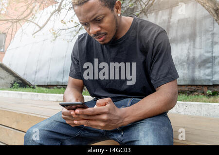 Dark-skinned man looking at mobile phone screen while sitting under tree Stock Photo