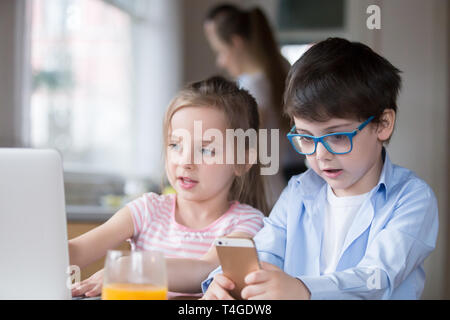 Children playing computer and smartphone while mother busy cooking breakfast Stock Photo