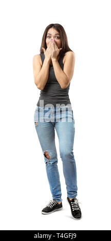 Young woman in gray top and blue jeans covering mouth and nose with her hands, weight on right foot, left knee lifted a bit, isolated on white Stock Photo