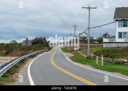 Winding road leads into the distance, among smallhouses, grass and bushes, and power line poles, Block Island, RI, USA Stock Photo