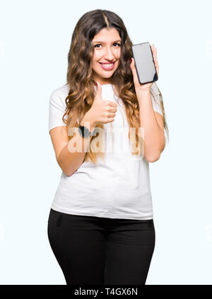 Young adult woman showing smartphone screen happy with big smile doing ok sign, thumb up with fingers, excellent sign Stock Photo