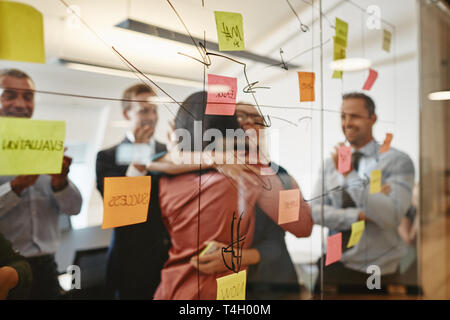 Two smiling young businesswomen hugging while brainstorming with sticky notes on a glass wall with coworrkers in a modern office Stock Photo