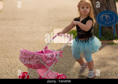 Happy child playing on grass. Beautiful little girl is playing with a doll in a stroller outdoors at sunset of the day Stock Photo