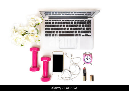 Women's workspace with laptop computer notebook, fitness equipment pink dumbbells, makeup cosmetics, mobile phone, headphones and white flower on white background. Stock Photo