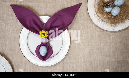 Beautiful Easter table setting with eggs, purple napkin Easter Bunny on white plates and on natural tablecloth background. Stock Photo
