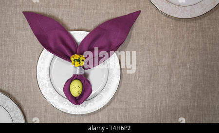 Beautiful Easter table setting with egg, purple napkin Easter Bunny on white plates and on natural tablecloth background. Stock Photo
