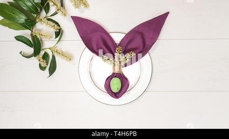 Beautiful Easter table setting with egg, purple napkin Easter Bunny, spring green tree branch on white wooden background. Stock Photo