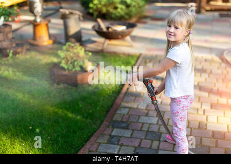 Little cute baby girl watering fresh green grass lawn mear house backyard on bright summer day. Child having fun playing with water hose sprinkler. Stock Photo