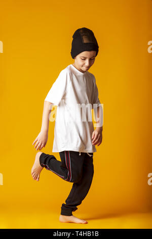 Cheerful little boy, dressed in sweatpants and a white T-shirt, actively dancing, standing on one leg. yellow background Stock Photo