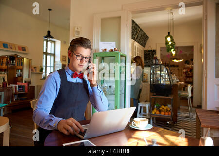 Shot of young man sitting at desk and talking with somebody on his mobile phone while working on laptop in his coffee store. Small business. Stock Photo