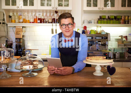 Portait shot of young coffee shop owner man wearing apron and holding digital tablet in his hand while standing behind shop counter and waiting for gu Stock Photo