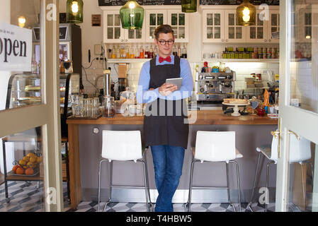 Portrait shot of smiling young coffee shop owner man standing in store and using digital tablet. Small business. Stock Photo