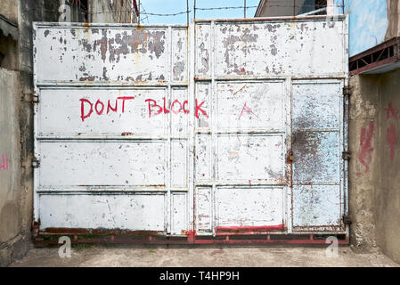 White colored old rusty metal entrance gate, closed and locked and secured with barbed wire. 'Don't Block' is written on the gate. Stock Photo