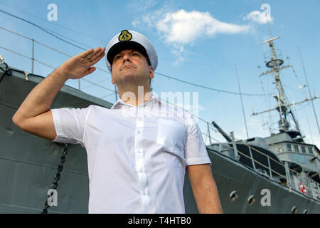 avy officer standing beside warship and do salute.The captain in white uniform stands under a battleship and saluting. Stock Photo