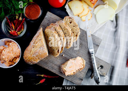 Close-up shot of Fresh sliced baguette  on wooden cutting board and spiced butter, tomatoes, cheese, bell pepper on dark table Stock Photo