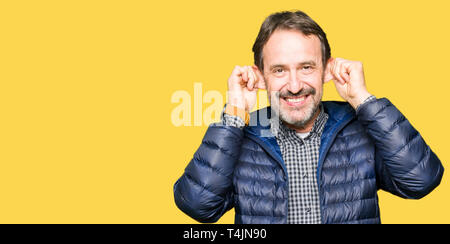 Middle age handsome man wearing winter coat Smiling pulling ears with fingers, funny gesture. Audition problem Stock Photo