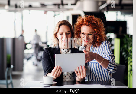 Two serious female business people with tablet sitting in an office, talking. Stock Photo