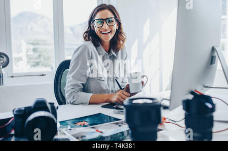 Smiling young woman photo editor with cup of coffee sitting at her desk. Female photographer in her office.
