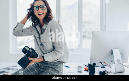Smiling female photographer with a professional camera sitting on her desk. Woman with dslr camera in office looking away and smiling. Stock Photo