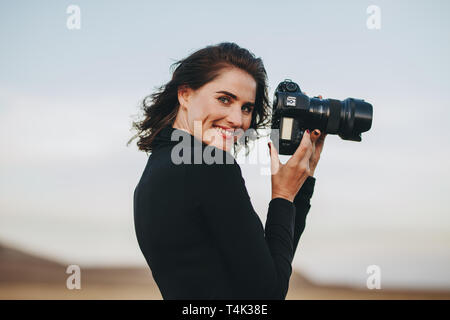 Positive photographer enjoying photo shooting outdoors. Young woman in casual holding a photo camera and smiling. Stock Photo