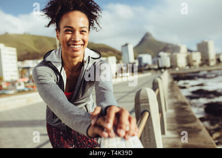 Woman runner exercising and smiling outdoors. Confident female runner in sportswear stretching on railing at seaside promenade. Stock Photo