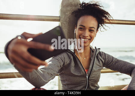 Smiling young woman runner in sportswear taking selfie with her mobile phone. Female relaxing and taking selfie after workout by the sea. Stock Photo