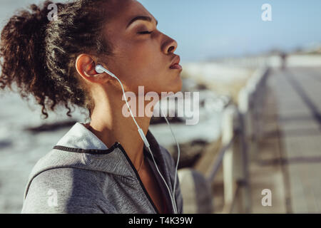 Tired and exhausted female runner resting after intense training session. Female with earphones taking a breather from physical training. Stock Photo