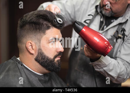 Close up view of master barber doing hairstyle with hairdryer at barber shop . Stock Photo