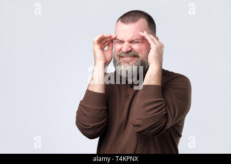 Mature man suffering from severe headache, pressing fingers to temples, closing eyes to relieve pain with helpless face expression Stock Photo