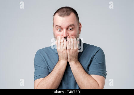Stressful terrified mature male with beard, covers mouth and looks down with eyes popped out down, being shocked. Negative emotions and frightening Stock Photo