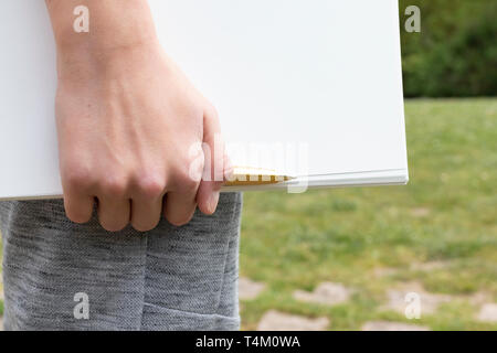 The boy's hand is holding a notepad with a yellow-black pen in the park against the background of green grass and trees. Clean white sheet. Going to w Stock Photo