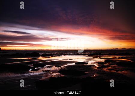 Sky with deep hanging storm clouds and wet sludge during low tide swathed in yellow and red bright light during sunset on tropical island Ko Lanta, Th Stock Photo