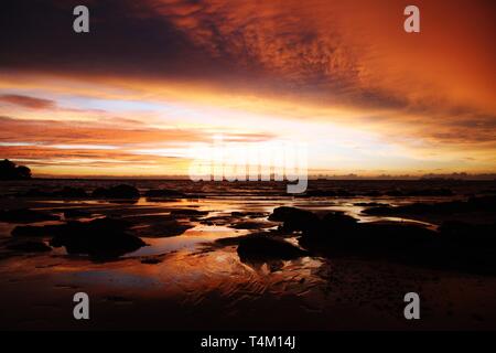 Sky with deep hanging storm clouds and wet sludge during low tide swathed in yellow and red bright light during sunset on tropical island Ko Lanta, Th Stock Photo
