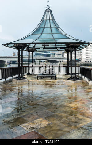 A couple embracing and sitting on a bench in a shelter on South Bank overlooking the River Thames in London. Stock Photo